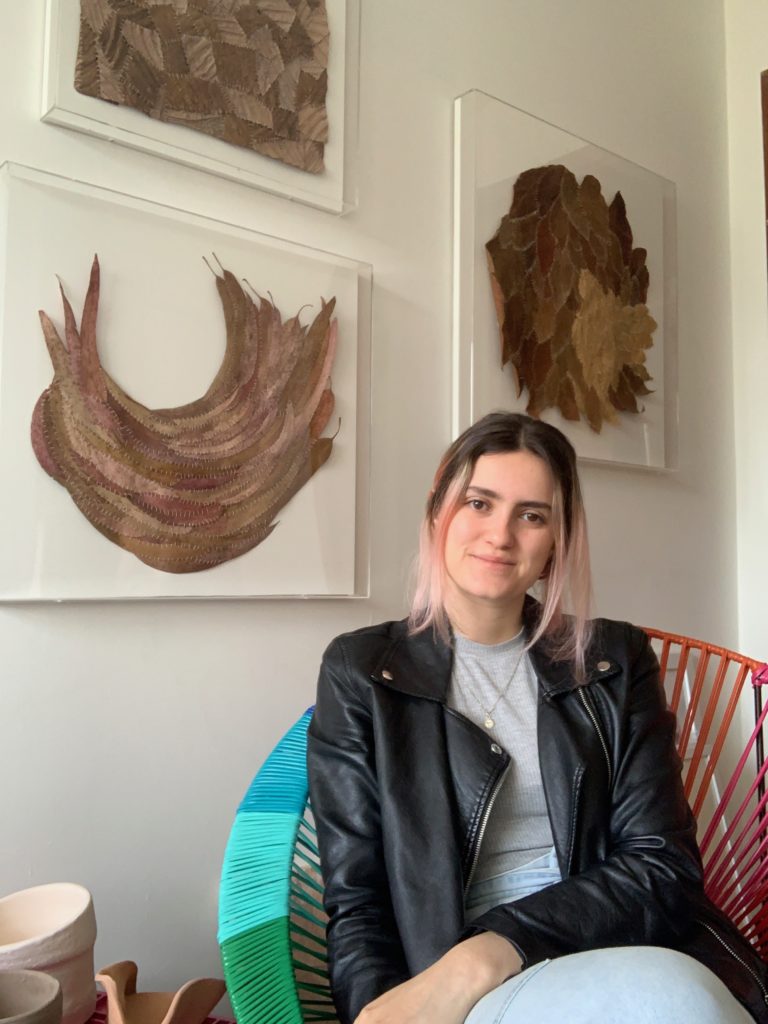 Porträt der jungen Künstlerin Helena Ospina Lizarralde, die auf einem Stuhl in einem Wohnzimmer mit Gemälden an der Wand hinter ihr sitzt und eine Lederjacke und ein graues T-Shirt trägt